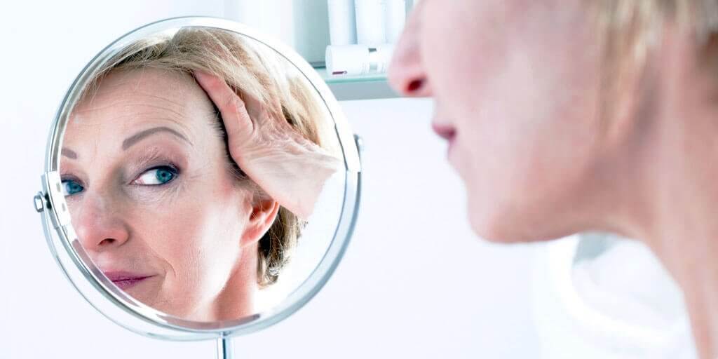Cosmetic-eye-at-a-glance-1024x512 A 40+ woman looking at her face in a mirror.