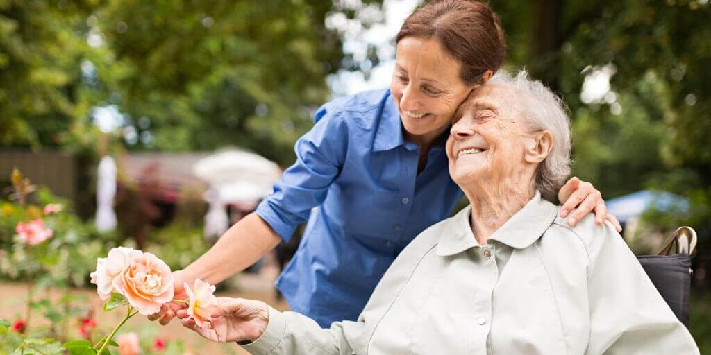 what-is-neuro-ophthalmology A disabled senior woman in a wheelchair touching flowers with the help of a younger woman caregiver.